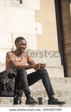Full length portrait happy young African American man sitting on steps outside with bag and cellphone