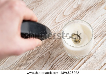 Using milk frother device in glass, closeup. Milk handheld mixer. Glass of milk on wooden table. High quality photo Royalty-Free Stock Photo #1896825292