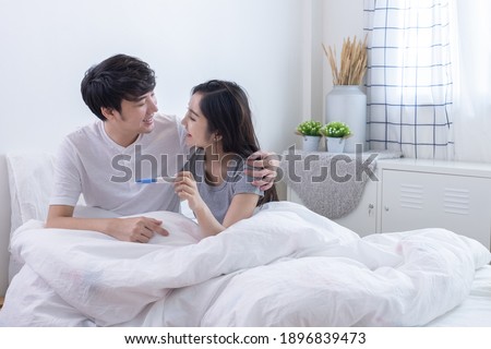 Happy young Asian couple looking and smiling together and wife holding pregnancy test in hand on bed in white bedroom.