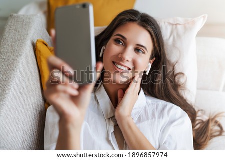 Close-up portrait. Woman is lying on couch, relaxing, using smartphone for text messages, exchanging photos, chatting with friends, checking emails, watching videos. Brunette girl smiling.