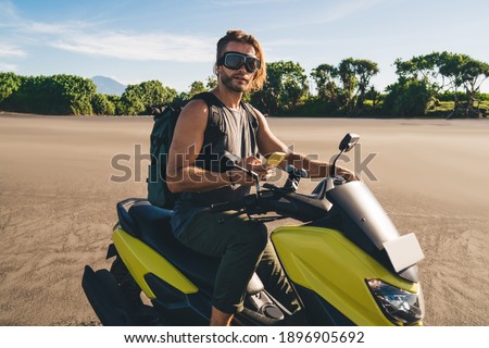 Side view of self esteem young stylish guy in casual clothes and goggles sitting on yellow motorbike with smartphone in hand while spending summer day on sandy beach
