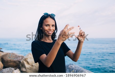 Cheerful blogger smiling at camera while using smartphone application for clicking photo images at coastline, happy female millennial holding cellular gadget and posing during summer vacations