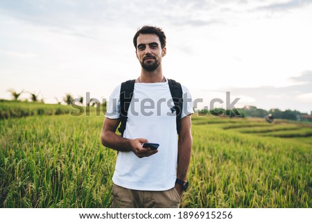 Bearded male tourist with dark hair wearing white shirt and backpack standing against green crops plantation and using mobile phone
