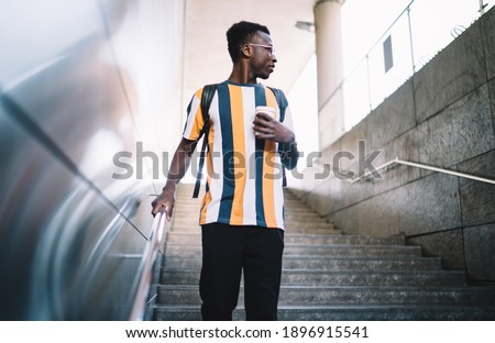 Pensive male tourist with caffeine beverage in hand standing at urban stairs and looking away pondering on solo travelling, African American hipster guy with takeaway coffee to go thinking in city Royalty-Free Stock Photo #1896915541