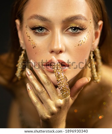 Stylish girl looks at camera. Gold ring on finger, earrings rings. Beautiful face, blue eyes, blonde hair, golden glamorous makeup, gold paint on body, skin, hands. Portrait of fashion model woman Royalty-Free Stock Photo #1896930340