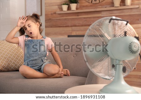 Little girl enjoying air flow from fan on sofa in living room. Summer heat Royalty-Free Stock Photo #1896931108