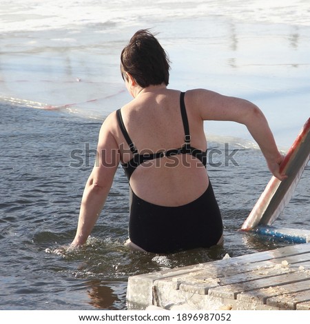 Winter swimming sport, a mature Russian woman in a black swimsuit enters the ice hole water on a Sunny frosty winter day, healthy lifestyle