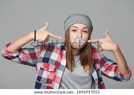 Cheeky teen girl blowing bubblegum and pointing at it, over grey background, studio portrait, isolated