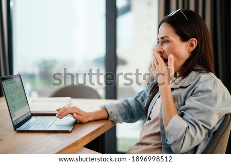 Attractive tired young smart woman working on laptop computer while spending time at home in the kitchen