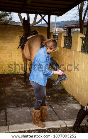 Riding woman dressed in cowboy clothes cleaning her riding boots with her horse tied to the bottom of the stable prepared with the saddle