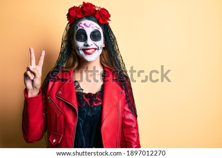 Woman wearing day of the dead costume over yellow smiling with happy face winking at the camera doing victory sign. number two. 