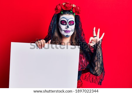 Young woman wearing day of the dead costume holding blank empty banner doing ok sign with fingers, smiling friendly gesturing excellent symbol 