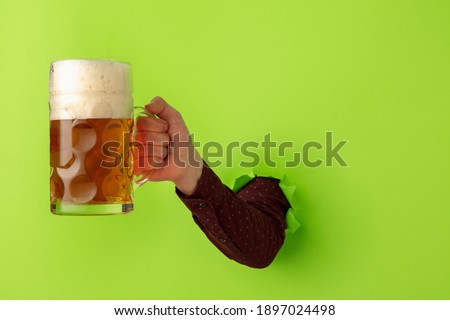 Close-up male hand with beer mug breaks through green paper background.