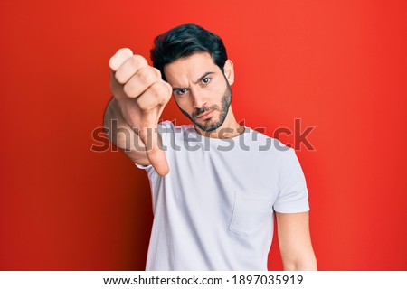 Young hispanic man wearing casual white tshirt looking unhappy and angry showing rejection and negative with thumbs down gesture. bad expression. 