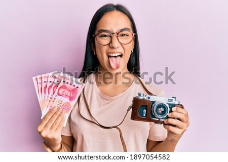 Young asian woman holding vintage camera and 100 chinese yuan sticking tongue out happy with funny expression. 