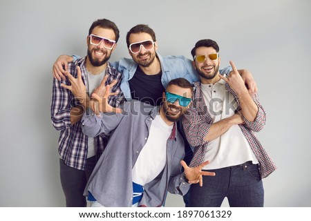 Happy best friends having fun while hanging out together. Group of 4 young men in sunglasses smiling, looking at camera and posing for funny photo in studio with light gray background