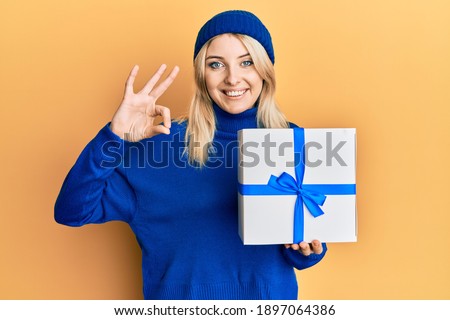 Young caucasian woman holding gift doing ok sign with fingers, smiling friendly gesturing excellent symbol 