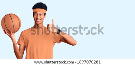 Young african american man holding basketball ball smiling happy and positive, thumb up doing excellent and approval sign 