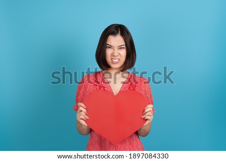 close-up angry, furious young Asian woman in a red dress holding a large red paper heart and baring her teeth, isolated on a blue background.