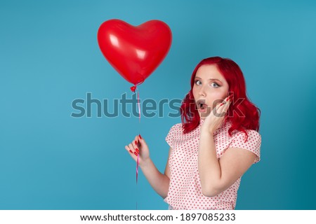 close up portrait of a surprised astonished young woman with red hair with open mouth holding a red flying balloon in the shape of a heart, isolated on a blue background.