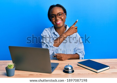 Young african woman working at hotel reception using laptop with a big smile on face, pointing with hand and finger to the side looking at the camera. 