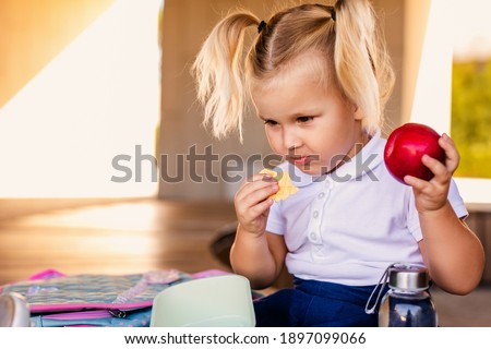 Cute smiling little schoolgirl holding a hamburger and orange juice outdoor - Back to school.
