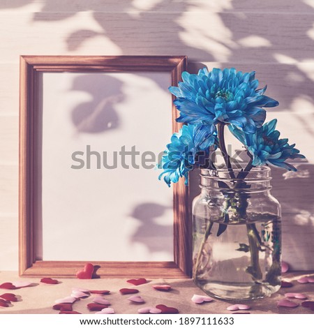 Empty wooden photo frame, blue chrysanthemums in vases, shapes in the form of hearts are on a light wooden background. Floral template for valentine's day, mother's day. Copy space.