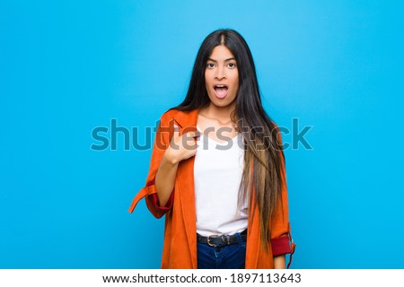 young pretty latin woman looking shocked and surprised with mouth wide open, pointing to self against flat wall