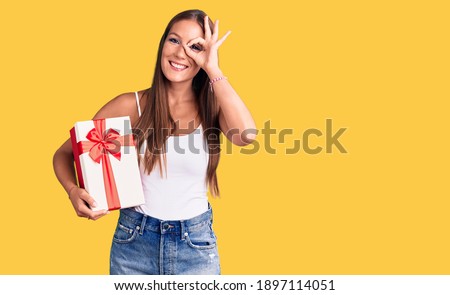 Young beautiful hispanic woman holding gift smiling happy doing ok sign with hand on eye looking through fingers 