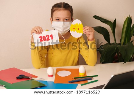 Girl with mask holding handmade easter egg card from decorative paper  during covid-19 quarantine. Children's DIY concept. Easter decoration or greeting card. Medical mask with 2021 text.