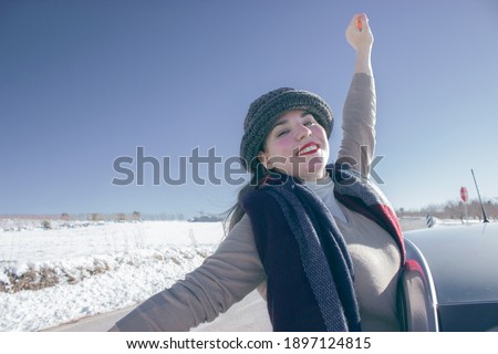 Woman is wearing hat and checkered scarf hanging out of the car window traveling on winter forest. Hipster girl admire snowy road. Traveler rest among stunning winter landscape.