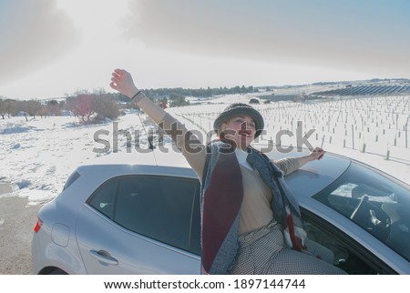 Beautiful woman traveler wearing hat and scarf lean out of the car window and enjoying of winter road and forest. Young hipster traveling by car in winter holidays.Winter Outdoor 2021.