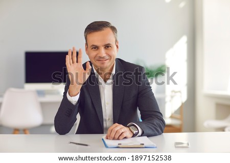 Happy smiling man in suit sitting at office desk, looking at camera and waving hand saying hello during video call. Webcam portrait of business coach, CEO, financial consultant or university professor Royalty-Free Stock Photo #1897152538