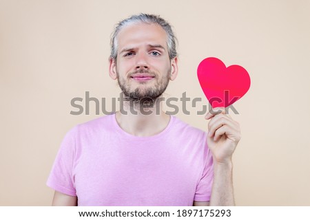 Cute smiling boy is holding red decorative sign of heart. Young man cupid in pink t-shirt is expressing sympathy, feelings. Happy St. Valentine's Day. Fall in love. Like concept.