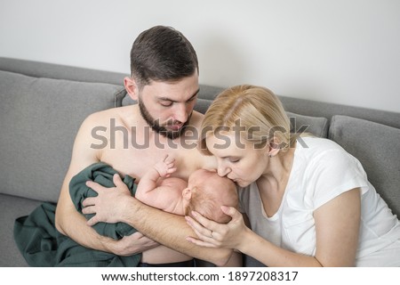 Parents hug a crying newborn baby at home. Dad holds his newborn son trapped in a trendy green muslin diaper skin to skin. Mom gently kisses the screaming baby. Baby care concept, first month of life.