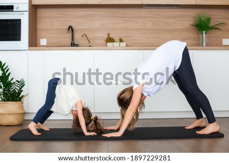 Woman and her little daughter are standing in a downward-facing dog pose while doing yoga together at home