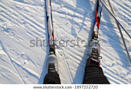 Feet of a man in ski boots stand on skis in the snow. Ski poles lie on the snow. The view from the top. Without a face. Sports, tourism. Outdoor activities in winter.
