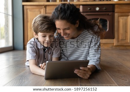 Smiling loving young Caucasian mom lying on warm floor using browsing tablet gadget with small son. Happy mother relax at home look at pad screen play online game or watch video with little boy child. Royalty-Free Stock Photo #1897256152
