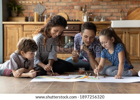 Happy young Caucasian family with two small kids sit on floor in cozy kitchen paint with colors together. Smiling loving parents relax play have fun drawing with little preschooler children at home.