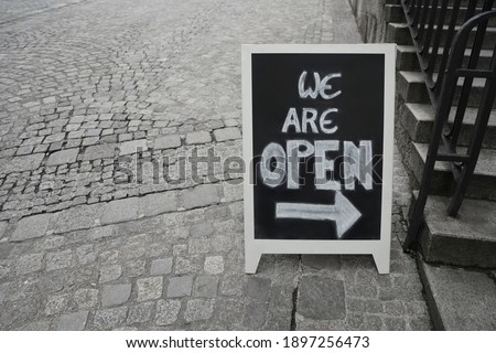 we are open - sign on a black chalkboard written in capital white letters.  The board stands on an empty street pavement with space. Stoned stairs with iron railings. Linz, Germany.