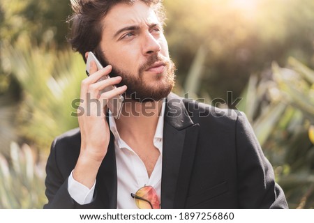 Handsome bearded young man in suit talking at the phone outdoors.