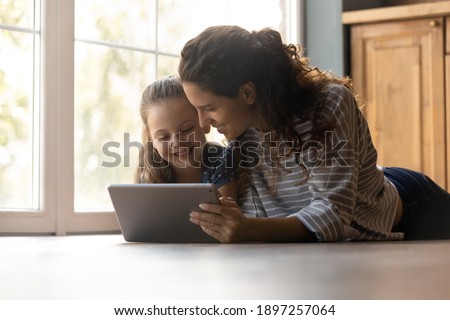Caring young mom and little daughter lying on warm home floor have fun studying learning using modern pad gadget. Happy mother and small girl child look at tablet screen watching video online. Royalty-Free Stock Photo #1897257064