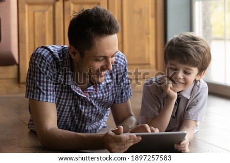 Caring young Caucasian father lying on warm floor relax have fun using tablet with little son. Happy dad and 6s small boy child rest at home look at modern pad screen browse or watch video together.