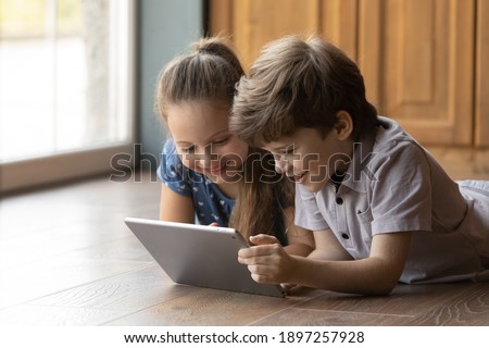 Close up of smiling small Caucasian children boy and girl lying on warm floor at home look at tablet screen playing game online. Happy two little kids have fun using modern pad gadget together.