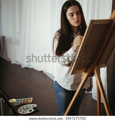 Art, hobby, DIY concept. Young concentrated woman do easel drawing, holding brush in hand. Illustrator or artist at work. Creative home leisure concept