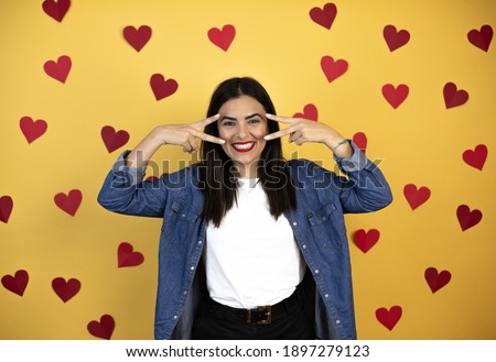Young caucasian woman over yellow background with red hearts Doing peace symbol with fingers over face, smiling cheerful showing victory