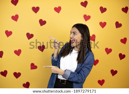 Young caucasian woman over yellow background with red hearts surprised and pointing her fingers side