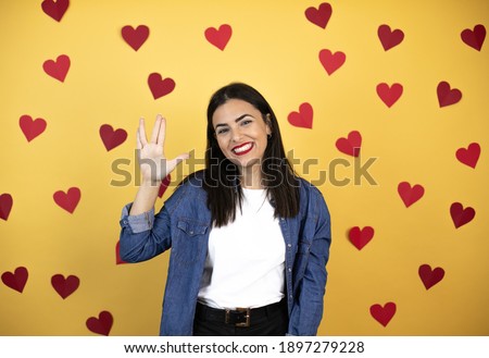 Young caucasian woman over yellow background with red hearts doing hand symbol