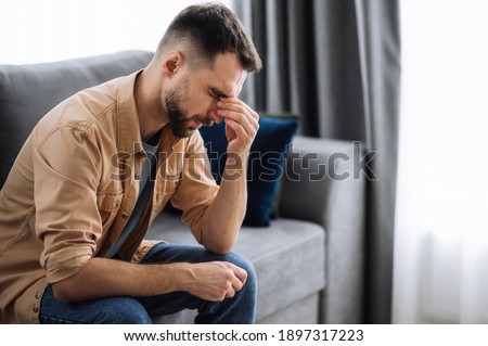 Tired guy sitting on the sofa at home, having a terrible headache. Exhausted caucasian male massage the eyes, feeling unhealthy, catch a cold or having flu, upset about illness Royalty-Free Stock Photo #1897317223
