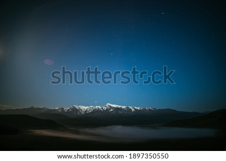 Atmospheric night landscape with dense fog and great snowy mountain ridge under starry sky. Alpine scenery with thick fog and big mountain range in starry night. Snow pinnacle under star night sky.